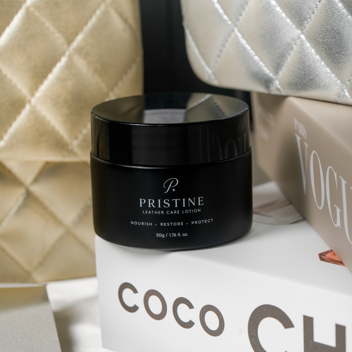 PRISTINE Leather Care Lotion on stacked fashion books with cream and silver quilted handbags.