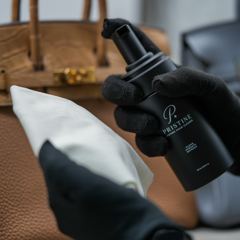 Black-gloved hands holding the PRISTINE Foam Cleaner pump bottle and a white cloth beside a tan handbag.