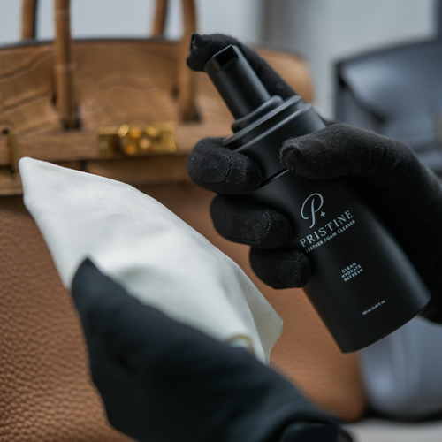 Black-gloved hands holding the PRISTINE Foam Cleaner pump bottle and a white cloth beside a tan handbag.