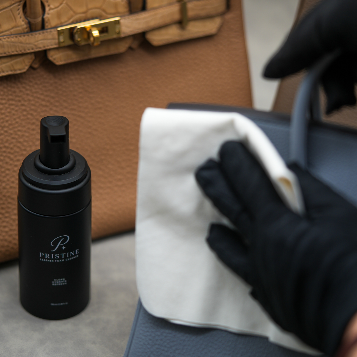 Black-gloved hand gently wiping a blue leather handbag with a white cloth beside PRISTINE Foam Cleaner.